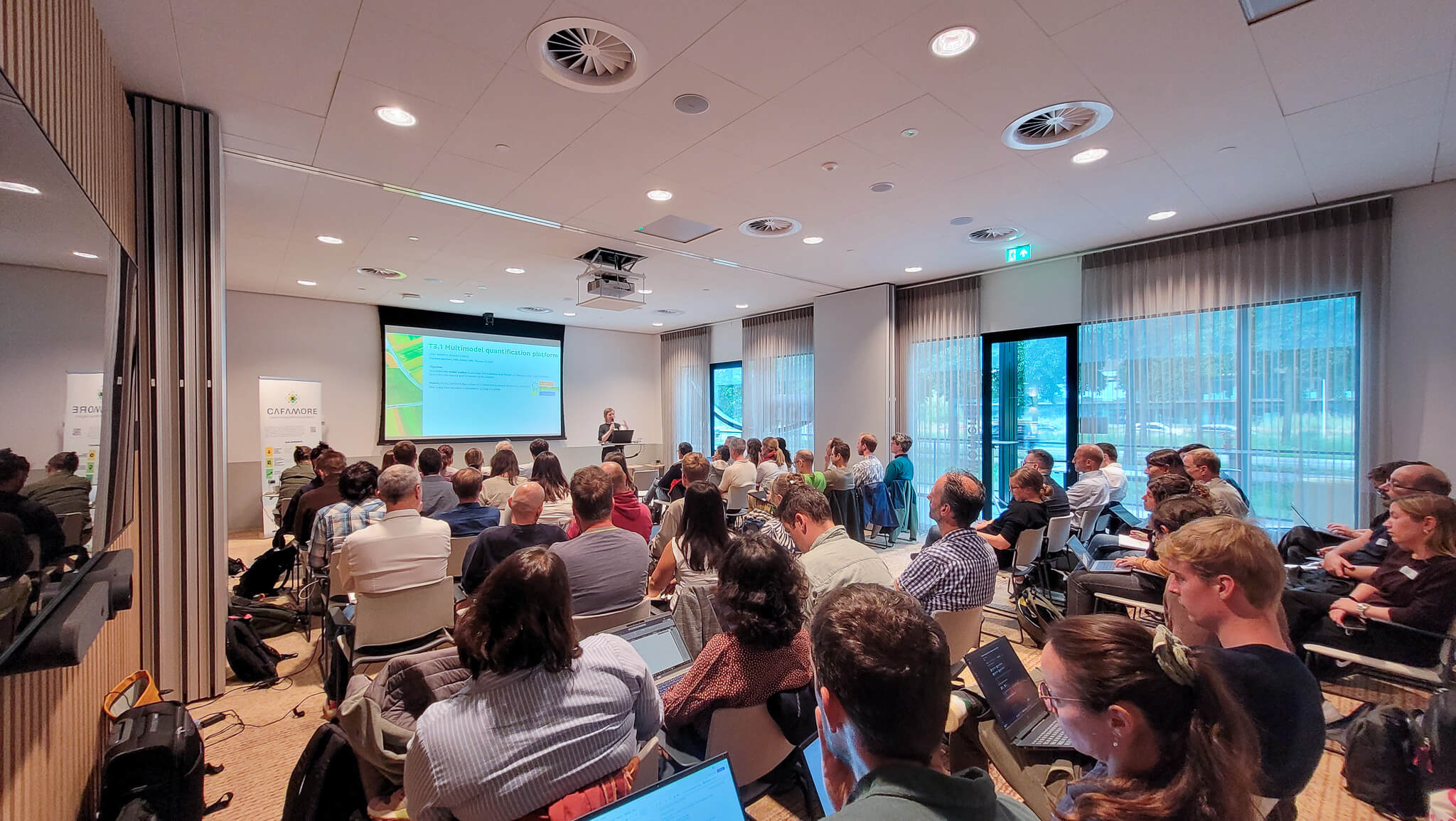 A large group of people seated in a room, focused on a presentation displayed on a projector screen.